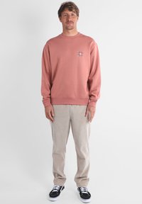 Pull couleur rose poudré, mélange de coton, col rond, poignets côtelés, petite broderie florale multicolore sur la poitrine, associé à un pantalon en velours côtelé beige.