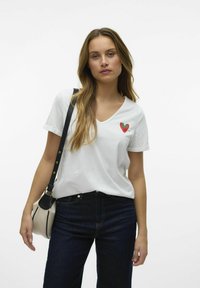 T-shirt blanc à col en V avec un petit motif de cœur rouge et vert. Associé à un jean bleu foncé et un sac à bandoulière noir. Texture lisse.