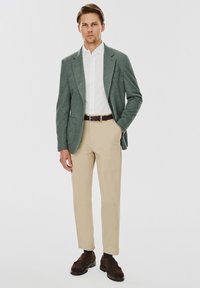 Jasnozielona marynarka na białej koszuli, zestawiona z beżowymi spodniami. Ciemnobrązowe buty uzupełniają strój. Gładki materiał i dopasowany krój.