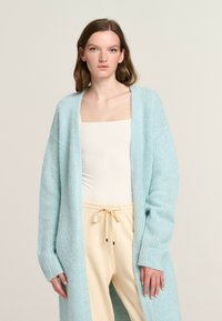 Light blue knitted cardigan with oversized sleeves, open front, and ribbed texture, worn over a cream tank and beige drawstring pants.