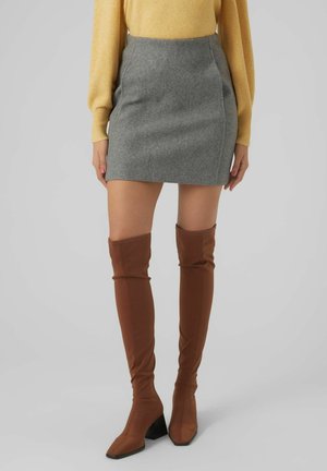 Kobieta ubrana w szarą mini spódnicę, żółtą bluzkę z długim rękawem oraz brązowe kozaki za kolano na obcasie, stojąca na jednolitym tle.