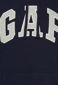 Sweat à capuche bleu marine avec de grandes lettres grises "GAP" sur la poitrine et deux cordons foncés suspendus à la capuche.
