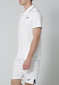 Biała koszulka polo Nike z czarnym logo, dopasowana, z krótkimi rękawami i kontrastowym wykończeniem przy kołnierzyku. Noszona z pasującymi białymi szortami.