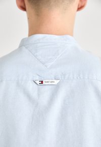 Ljusblå skjorta med krage och slät textur, med en etikett som har "TOMMY JEANS" nära baksidan av halsringningen och en triangulär sömdetalj.