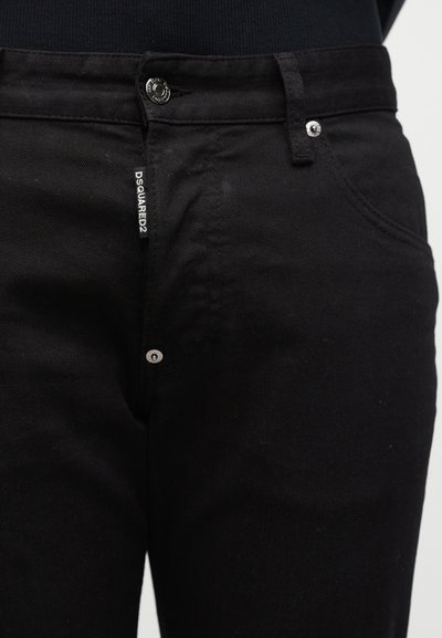 Κοντινό πλάνο μπροστινής όψης μαύρων τζιν με μεταλλικό κουμπί, πριτσίνια και μια μικρή ετικέτα DSQUARED2 κοντά στην περιοχή του φερμουάρ.