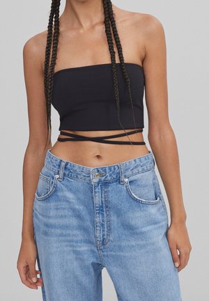 Femme avec de longues tresses portant un crop top noir sans bretelles avec des liens enroulés et un jean taille haute bleu clair sur fond gris.