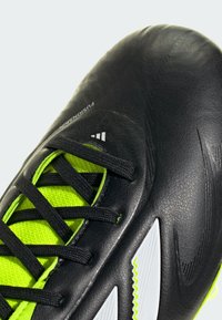 Zwarte voetbalschoenen met een gestructureerde synthetische bovenkant, groene accenten en zwarte veters. Bevat details in witte panelen en branding.