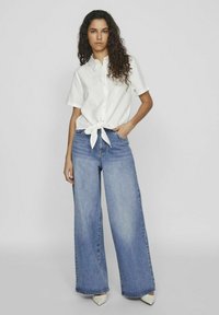 Camicia bianca con bottoni e davanti annodato, abbinata a jeans in denim blu chiaro a gamba larga e scarpe beige a punta. Tessuto morbido, vestibilità comoda.