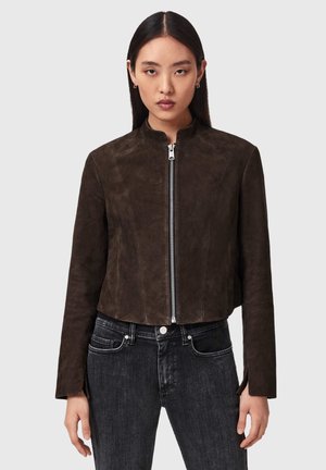 Vrouw draagt een donkergroen suède cropped jas met voorrits, gecombineerd met een high-waist zwarte spijkerbroek, staand tegen een effen achtergrond.