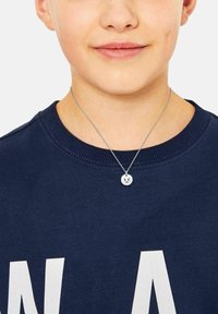 Collana d'argento con un pendente rotondo che presenta un design con una faccia arrabbiata, indossata sopra un felpa blu navy con lettere bianche.