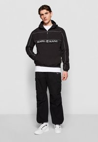 Sort vindjakke med logo på brystet, hvid piping, hætte og elastiske manchetter. Kombinationen er med sorte cargo-bukser og hvide sneakers.