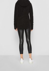 Musta fleece-huppari, jossa on huppu, yhdistettynä kiiltäviin mustiin legginsseihin ja valkoisiin lenkkareihin. Sileät pinnat ja istuva muotoilu nilkkapituisten legginsien kanssa.