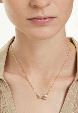 Guldhalsband med hänge i form av en oändlighetssymbol, prytt med små klara stenar och en central rund ädelsten, bärs på en bar hals.