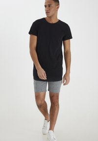 Sort bomulds T-shirt med opkradse ærmer, parret med grå ternede shorts. Modellen har hvide sneakers på og står foran en ensfarvet baggrund.