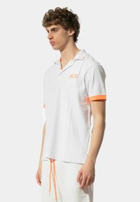Polo bianco con accenti arancioni sul colletto e sulle maniche. Presenta un piccolo logo arancione sul petto. Realizzato in un tessuto liscio.