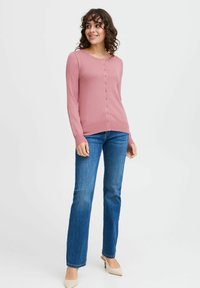 Gilet ajusté rose clair avec un col rond, patte de boutonnage à l'avant et manches longues, associé à un jean bleu et des talons de couleur crème.
