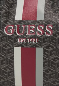Musta kassi geometrilise mustriga, millel on punane ja valge vertikaalne triip ning tugevate punaste tähtedega "GUESS EST. 1981". Tekstureeritud pind.