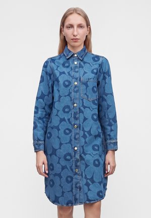 Kvinde iført en blå denimskjortekjole med store blomsterprint og guldknapper, stående foran en ensfarvet hvid baggrund.