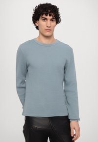 Šviesiai mėlynas ilgomis rankovėmis vaflinės tekstūros megztinis su grioveliais, apvaliu kaklu ir laisvu siluetu. Dėvimas su juodomis kelnėmis.