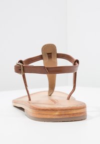 Hnědá kožená sandála s plochou dřevěnou podrážkou, strapcovým designem a nastavitelným přezkovým páskem na kotníku. Hladký povrch s minimálně viditelným šitím.