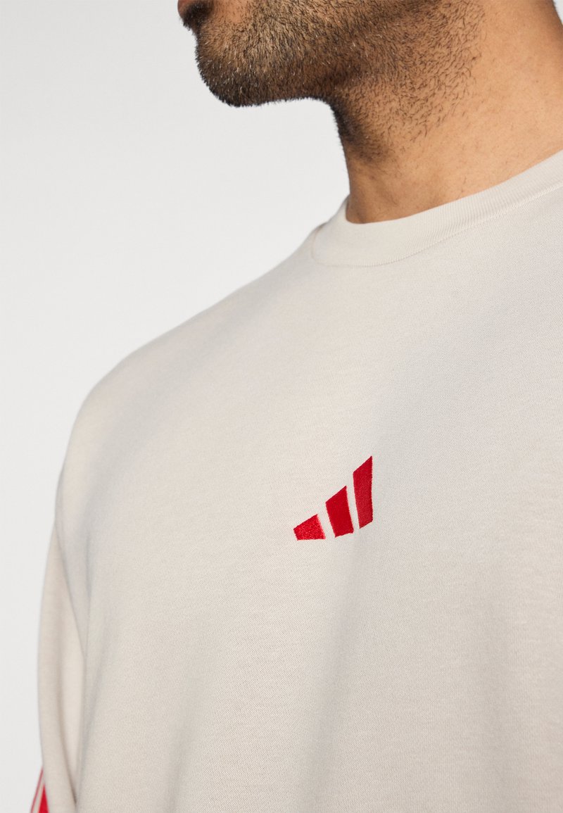 Close-up van een man die een lichtbeige shirt draagt met een klein rood geometrisch logo op de borst, waarbij een deel van zijn nek en baard zichtbaar is.
