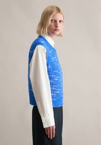 Niebieski sweter z dzianiny o teksturowym wzorze, noszony na białej koszuli z kołnierzykiem. Dolny brzeg jest prosty, a rękawy są bezrękawnikowe.