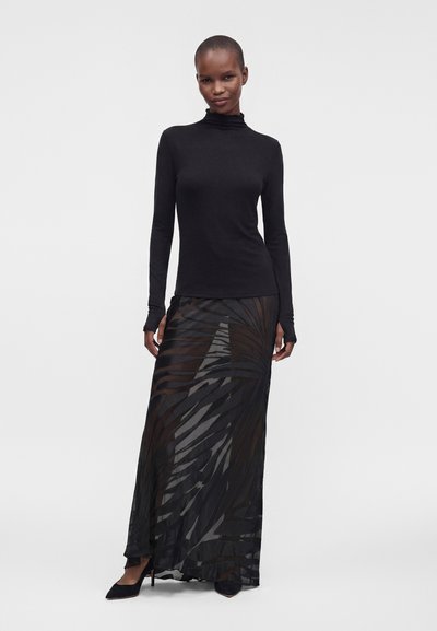 Femme debout, portant un haut à col roulé noir à manches longues et une jupe longue noire transparente à motifs avec des escarpins noirs pointus sur fond uni.