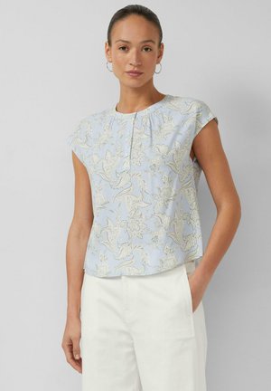 Vrouw draagt lichtblauw bloes met korte mouwen en witte broek, staat met één hand in haar zak tegen een effen grijze achtergrond.