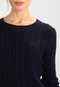 Marineblauwe gebreide trui met een gestructureerd kabelpatroon, ronde hals en lange mouwen. De stof lijkt zacht en warm.