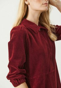 Veste en velours côtelé bordeaux avec un col, fermeture éclair et manches froncées. Texture côtelée et détails en motif vertical.