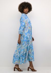 Maxi obleka iz svetlo modrega blaga s paisley vzorcem. Ima okrogel ovratnik, 3/4 rokave in lahkoten kroj. Rjave sandale z visokim peto.