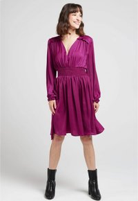 Robe violette mi-longue avec une taille smockée, des manches longues et un décolleté en V. Tissu lisse avec un léger éclat et une coupe décontractée.