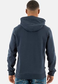 Navyblauwe hoodie van zacht materiaal met een grote capuchon, geribbelde mouwen en onderrand. Voorzien van een minimalistisch ontwerp en een label aan de onderkant links.