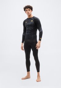 Νέος άνδρας όρθιος, ξυπόλυτος, φοράει μαύρο μακρυμάνικο συμπιεστικό τοπ με σχέδια και μαύρα συμπιεστικά κολάν, μπροστά σε απλό φόντο.