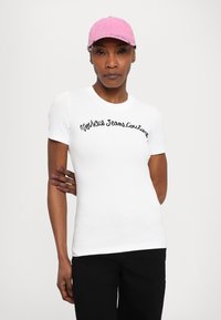 Balts īsās piedurknes T-krekls ar melnu izšuvumu "Northside Jeans Couture". Saskaņots ar rozā cepuri. Gluda materiāla, piegulošs dizains.