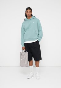 Sudadera de color teal claro con bolsillo y logo, pantalones cortos negros, mochila gris de Nike, calcetines blancos y zapatillas deportivas. Conjunto atlético casual con un ajuste relajado.