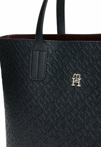 Zwarte textuur tote bag met een verhoogd geometrisch patroon. Beschikt over dubbele handvatten en een gouden logo accent. Gevoerd met bordeaux.