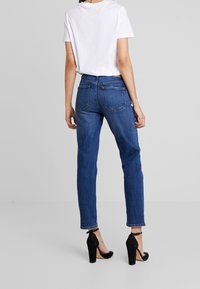Kvinna som står med ryggen mot oss, iklädd en vit t-shirt, blå jeans och svarta högklackade skor med ankelremmar mot en enfärgad grå bakgrund.