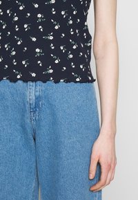 Top a canotta a coste blu navy con stampa floreale bianca e orlo scallop, abbinato a jeans a vita alta blu chiaro. Mano posata sui jeans.