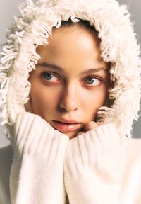 Biały sweter z dzianiny z grubym, puszystym kapturem. Tkanina jest miękka w dotyku i ma kudłatą teksturę. Twarz częściowo zasłonięta, ukazująca gładką skórę i jasne oczy.