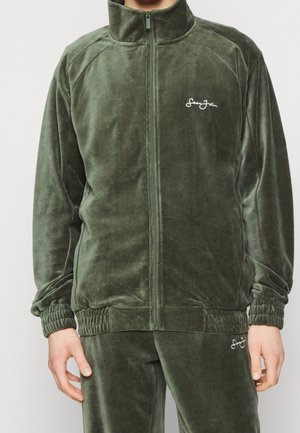 Personne portant un survêtement en velours vert olive avec veste zippée et pantalon assorti, tous deux avec de petits logos blancs brodés.