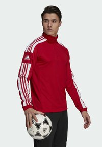 Red athletic jacket with white stripes on sleeves, zippered collar, and ribbed cuffs, worn by a person holding a black and white soccer ball.