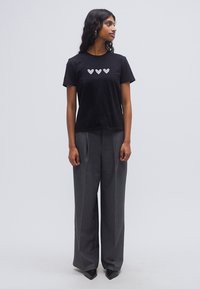 Μαύρο βαμβακερό T-shirt με τρία λευκά σχέδια καρδιών στο μπροστινό μέρος, συνδυασμένο με ψηλόμεσα γκρι παντελόνια με φαρδιά μπατζάκια και μυτερά μαύρα παπούτσια.