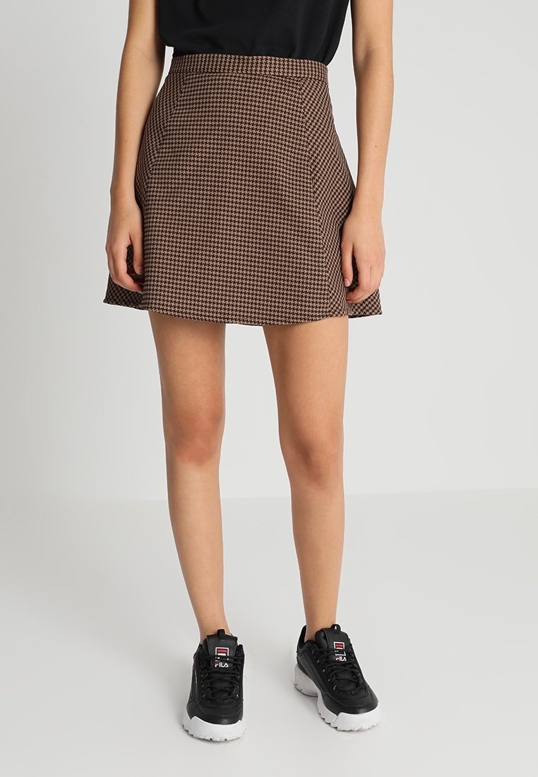 Brauner Hahnentrittmuster-Minirock mit hohem Bund, kombiniert mit einem schwarzen Oberteil und schwarzen Sneakers. Glattes Material mit einem leichten Schwung am Saum.