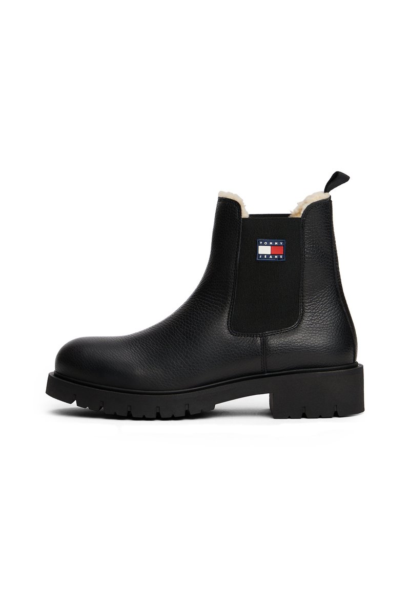 Bottine en cuir noire avec semelle épaisse, côtés élastiques, doublure en fourrure de mouton, et logo Tommy Jeans sur le côté extérieur.