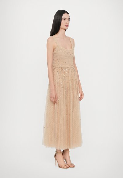 Abito in tulle beige con paillettes, corpetto aderente e gonna svasata. Presenta spalline arricciate e lunghezza a metà polpaccio. Indossato con tacchi alti nudi.