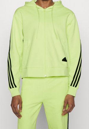 Person iført limegrøn hættetrøje og matchende bukser med sorte triple striber på ærmer og bukseben, stående mod en hvid baggrund.