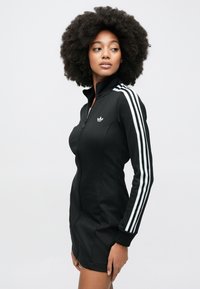 Fekete cipzáros ruha, fehér háromcsíkos mintával az ujjakon. Magas nyakkal és testhezálló szabással rendelkezik, sima, rugalmas anyagból készült.