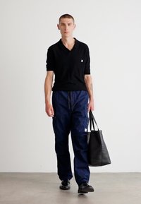 Black short-sleeve polo shirt, navy cargo pants with pockets, black leather tote bag, and black shoes, standing against a plain background.