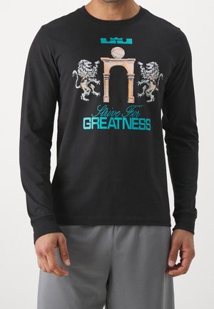 Chemise noire à manches longues avec deux lions flanquant une arche en pierre, un ballon de basket au-dessus et un texte turquoise indiquant « Strive for GREATNESS ».